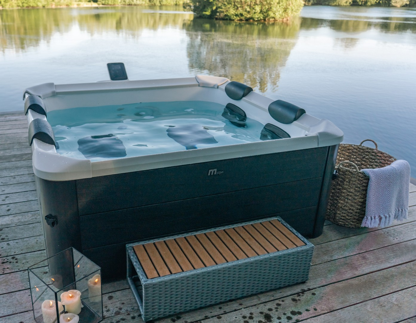 Ein Whirlpool mit Wasser steht auf einem Holzdeck. Im Hintergrund befindet sich ein See mit Bäumen. Eine kleine Stufe befindet sich vor der Wanne.