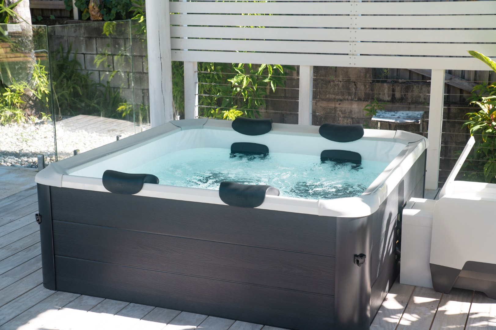 Ein quadratischer Whirlpool mit sprudelndem Wasser. Schwarze Kopfstützen sind innen. Er steht auf einer Holzterrasse, mit Zaun und Grün im Hintergrund.