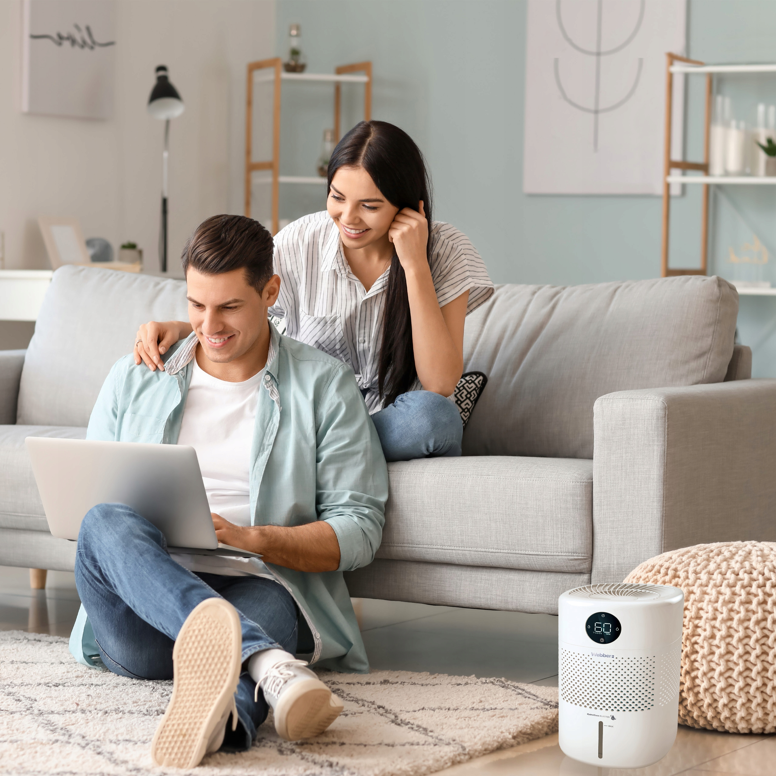 Para siedzi obok kanapy, używając laptopa. Osuszacz powietrza stoi na podłodze. Dywan jest na pierwszym planie.