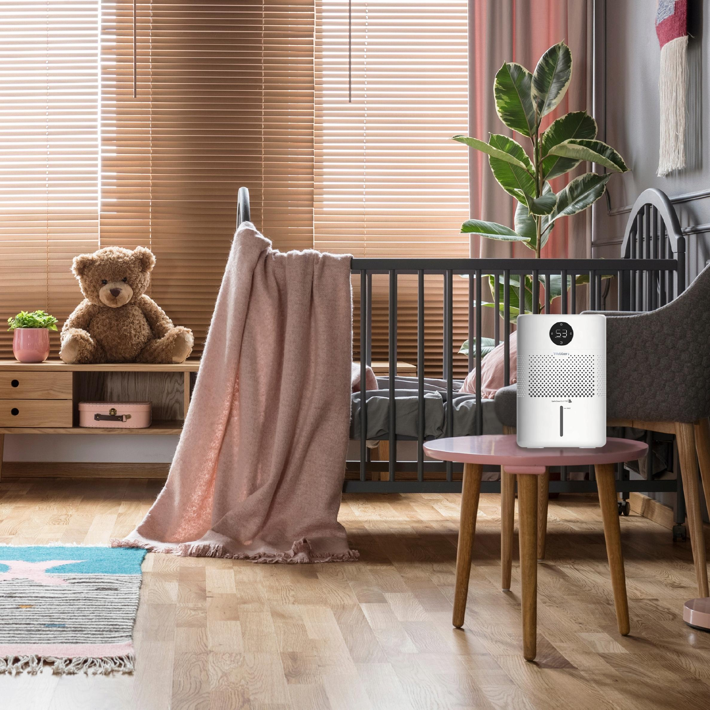 Sypialnia z łóżeczkiem, misiem i nawilżaczem na stole. Widoczne są również żaluzje i roślina.