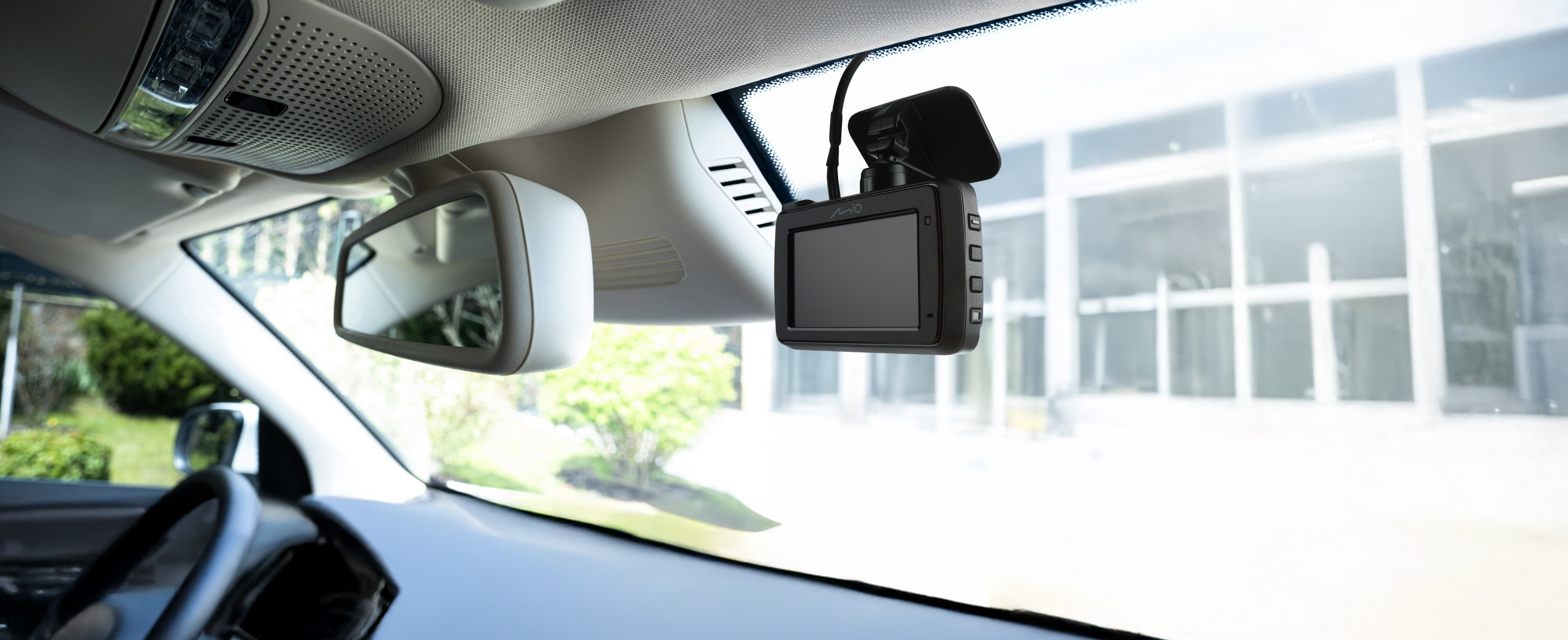 Interni auto. Una dashcam è appesa al soffitto. Specchietto retrovisore a sinistra. Le finestre riflettono un edificio con alberi.