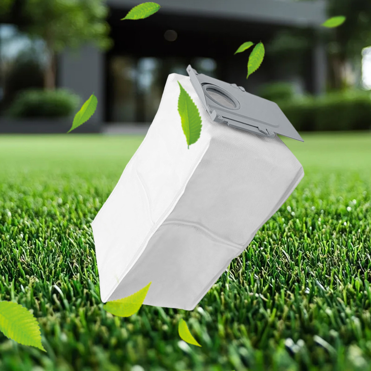 Biały worek do odkurzacza z szarą górą unoszący się nad zieloną trawą z zielonymi liśćmi w powietrzu i domem w tle.