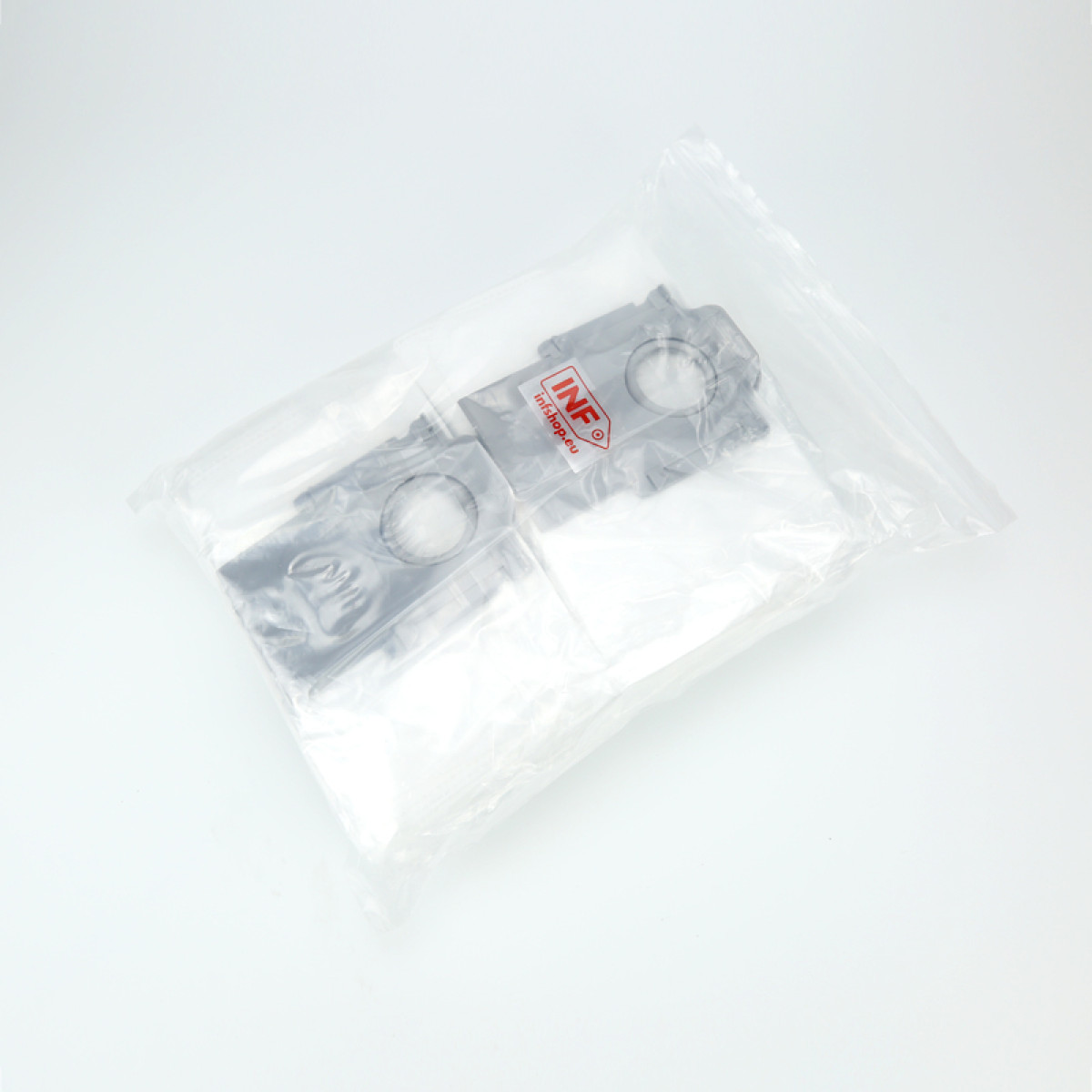 Zwei graue Kunststoffteile in einer transparenten Plastiktüte. Die Tüte liegt auf einer weißen Oberfläche. Die Teile haben kreisförmige Löcher.