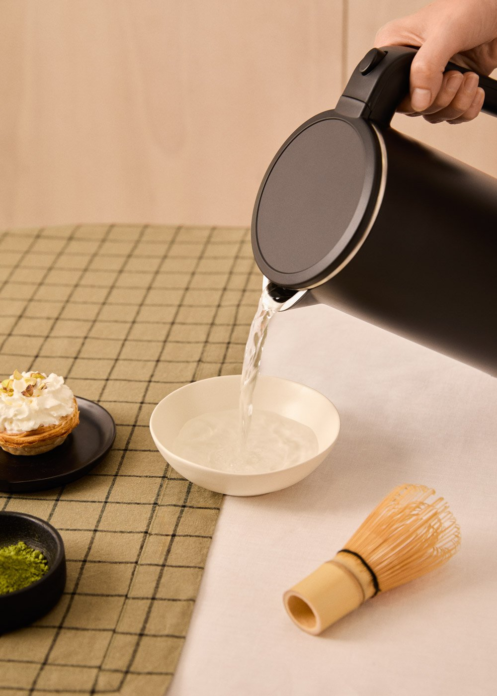 Una tetera negra vierte agua en un cuenco blanco. Una tarta, matcha y un batidor están sobre la mesa.