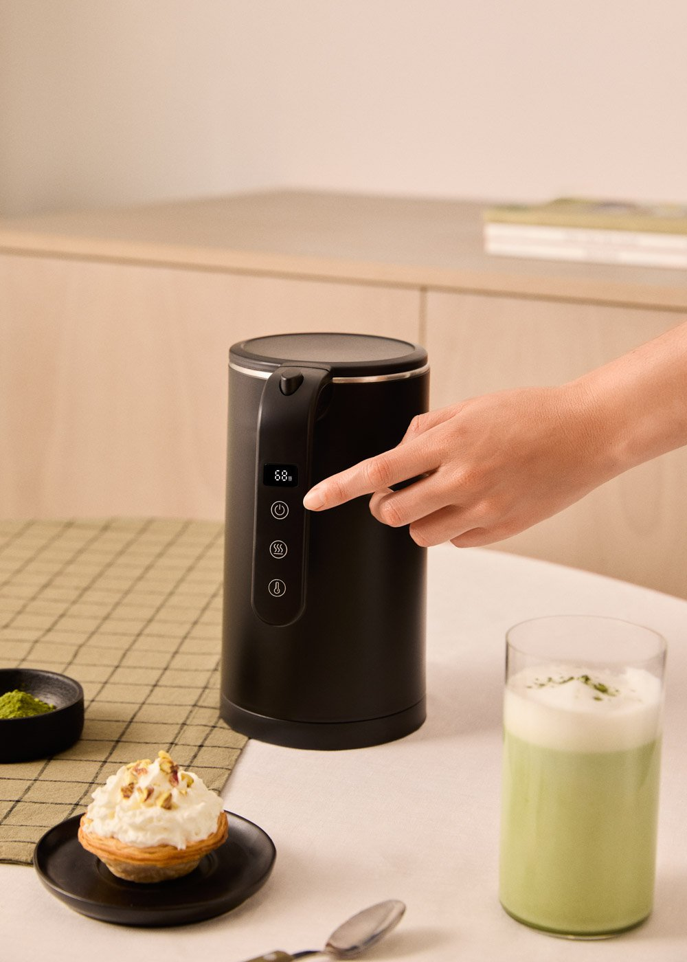 Una mano presiona un botón en una tetera eléctrica negra. Un latte matcha, una tarta y polvo de matcha están en la mesa.