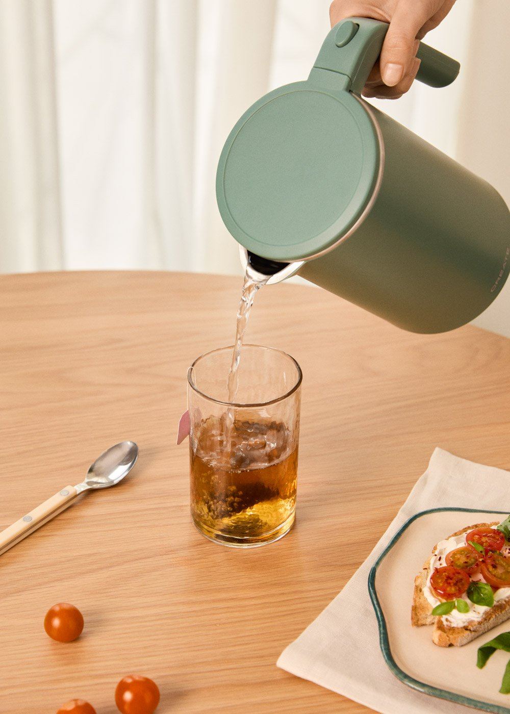 Una persona vierte agua de una tetera verde en un vaso de té. Tomates y un sándwich en la mesa.