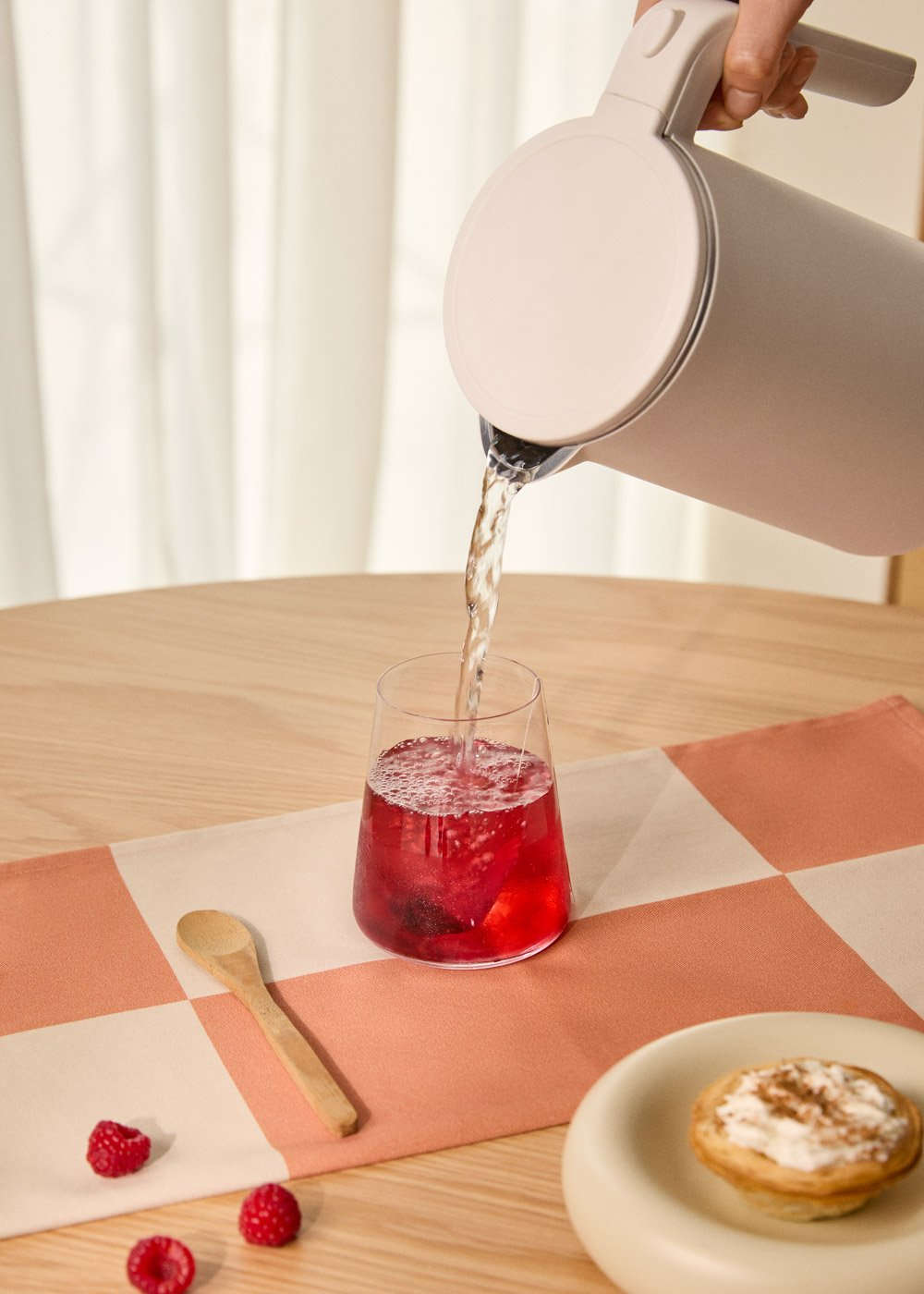 Agua vertiéndose en un vaso con líquido rojo. Frambuesas, una cuchara y un pastelito están sobre una mesa con un camino de mesa a cuadros.