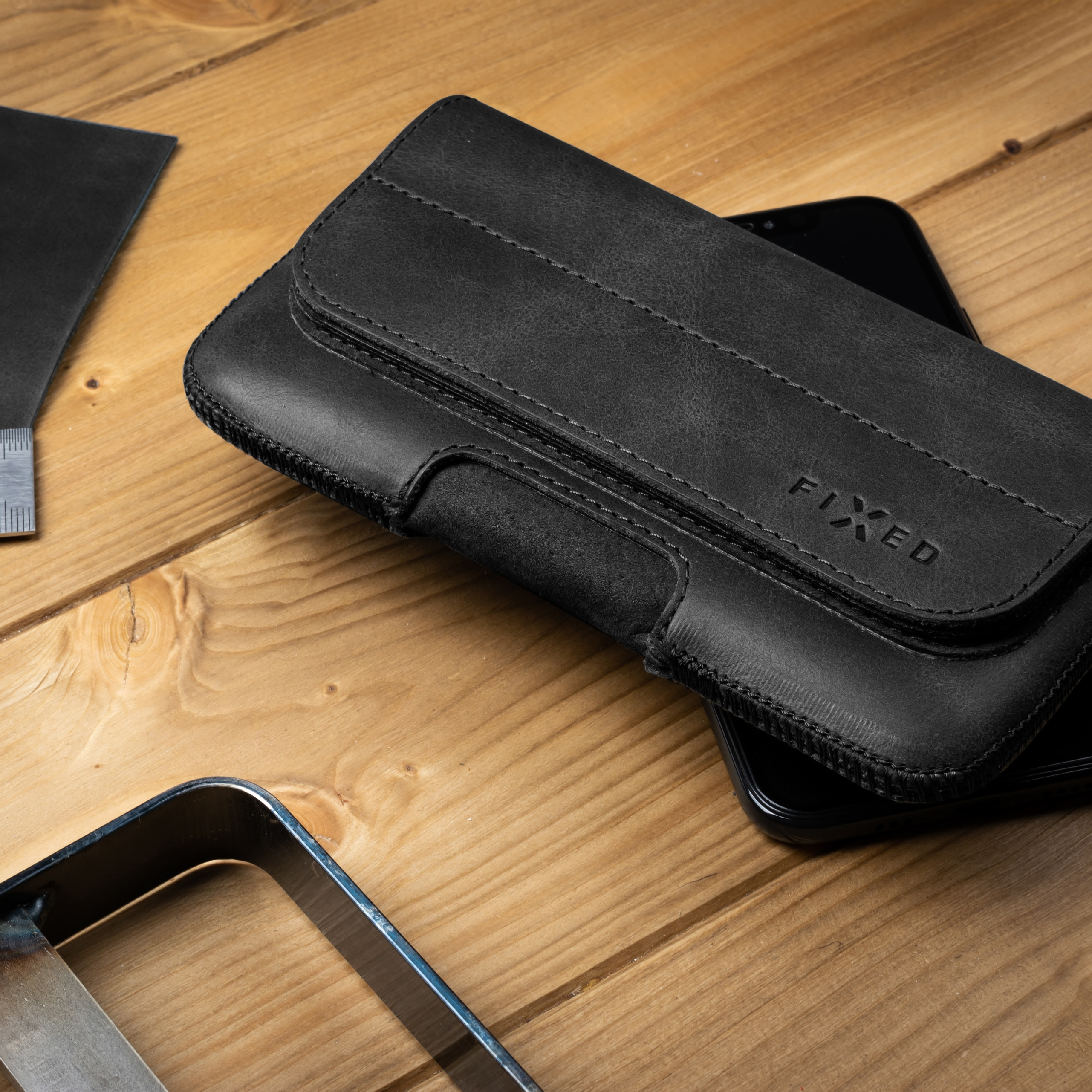Schwarze Leder-Handyhülle mit 'FIXED'-Logo auf Holzoberfläche mit Werkzeugen.