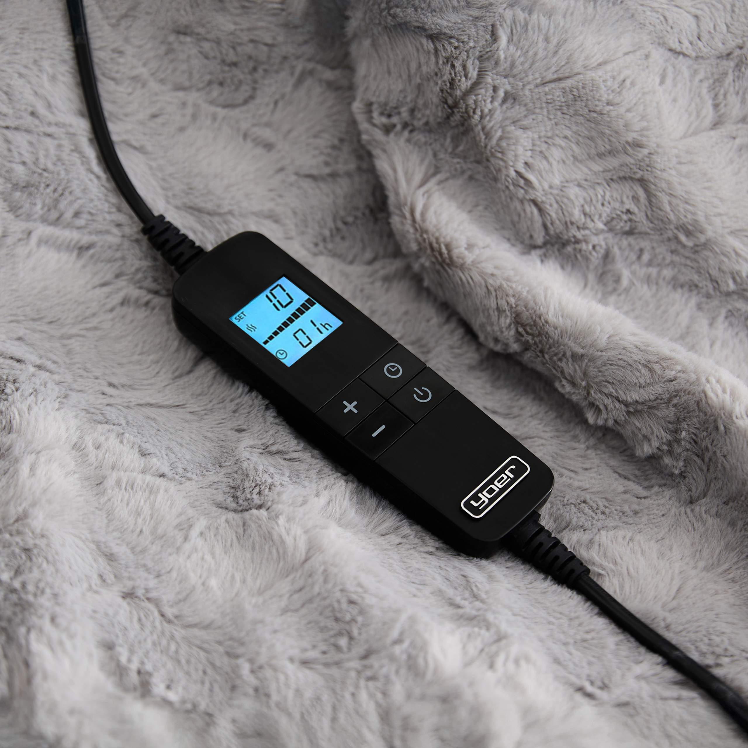 Czarny kontroler z ekranem, przyciskami i kablem zasilającym na puszystym, szarym kocu. Ekran pokazuje ustawienia.