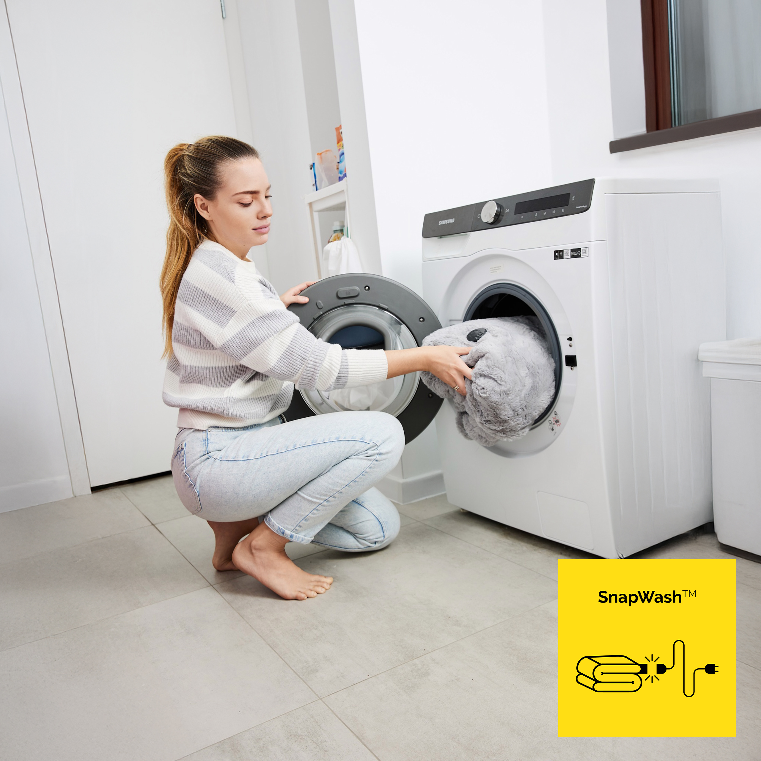 Kobieta wkłada ubrania do pralki. Jest boso. Pralka jest biała, a pokój ma szare płytki.