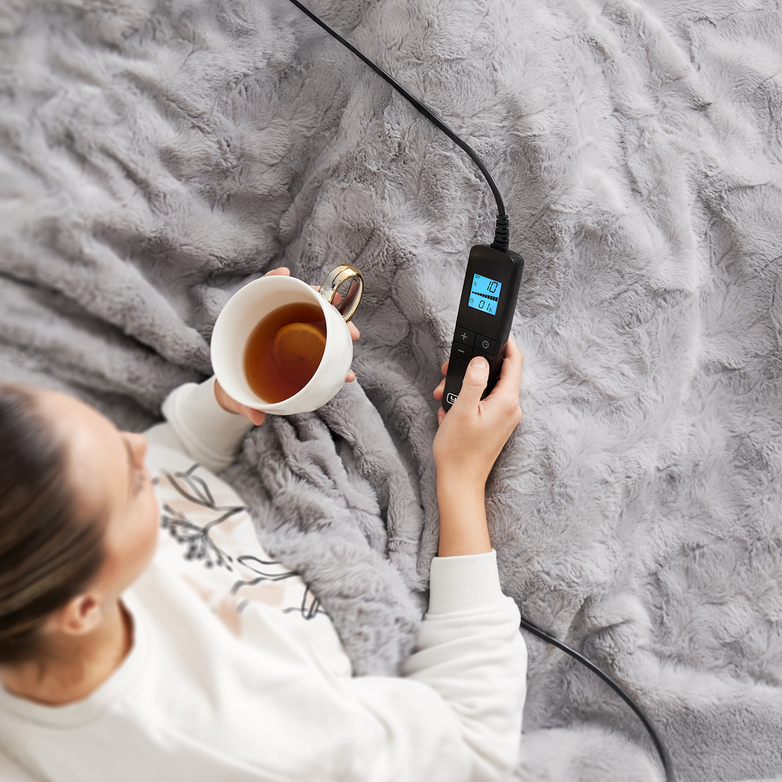 Osoba trzyma kubek herbaty i panel sterowania podgrzewanym kocem na szarym kocu.