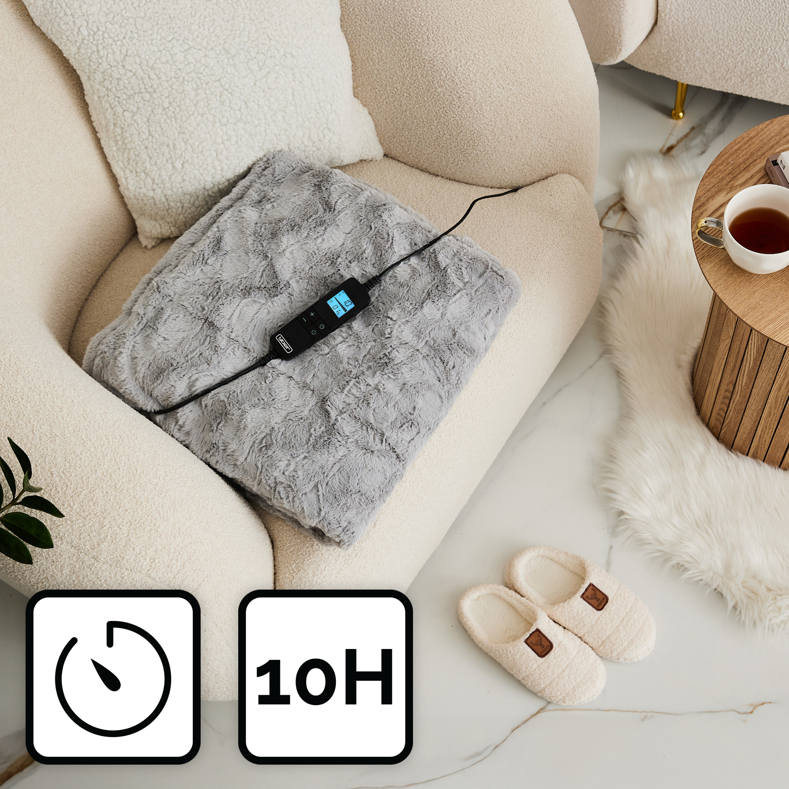 Szary koc grzewczy na beżowym krześle. Obok kremowe kapcie, biała poduszka i drewniany stolik z herbatą.