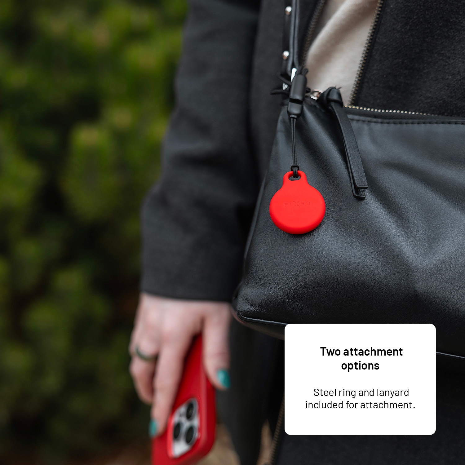 Eine Person hält ein rotes Telefon und eine schwarze Tasche mit einem roten Anhänger. Grünes Laub im Hintergrund.