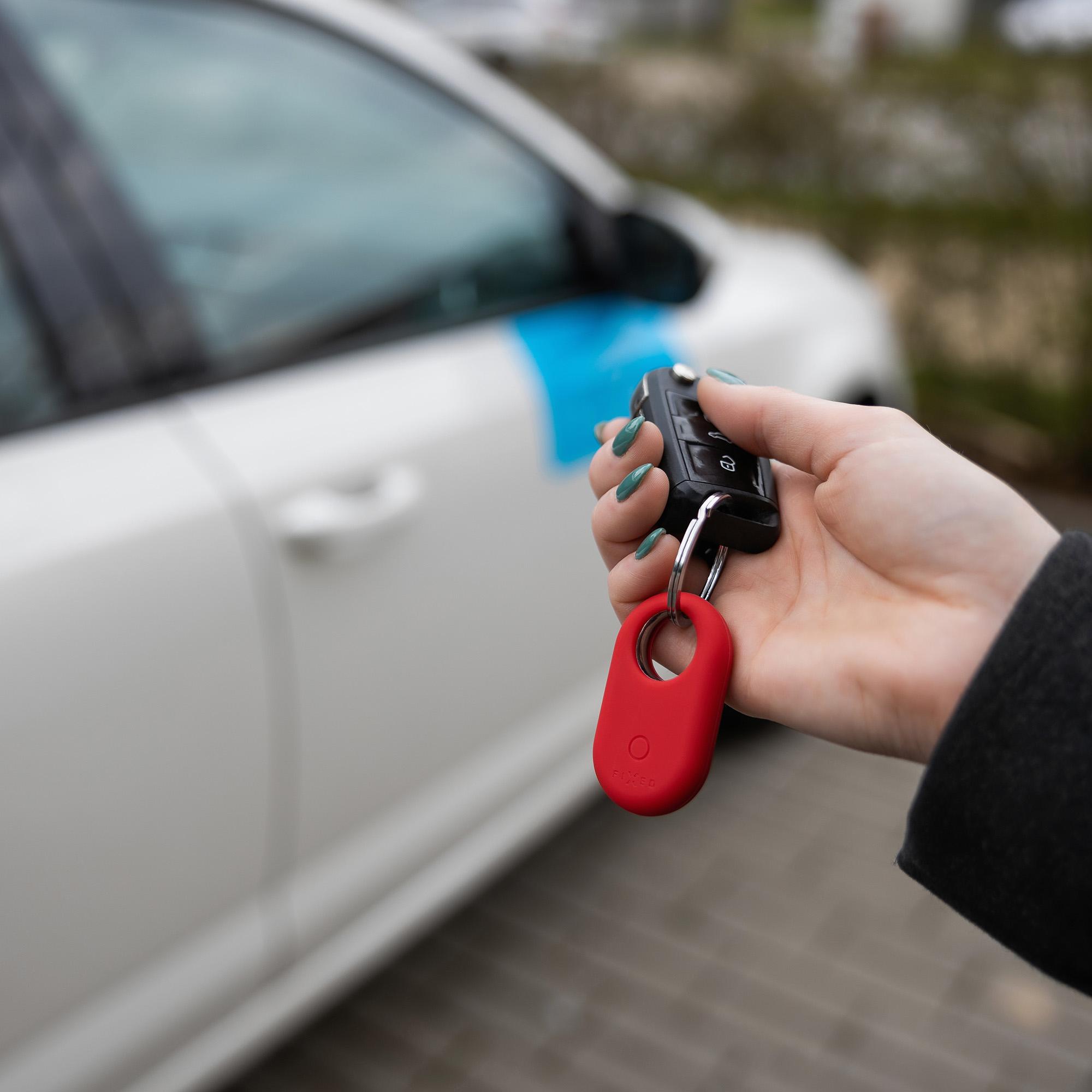 Eine Hand hält Autoschlüssel mit rotem Schlüsselanhänger neben einem weißen Auto. Grüner Nagellack ist sichtbar.