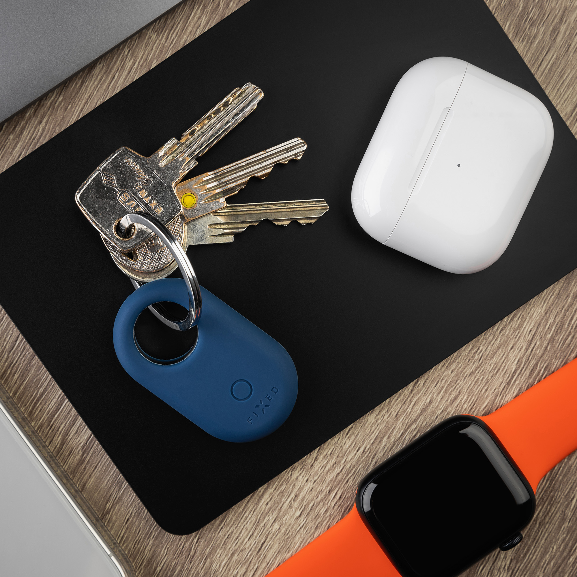 Schlüssel, blauer Schlüsselfinder, AirPods-Hülle und eine orangefarbene Smartwatch auf Holz und schwarzem Pad.