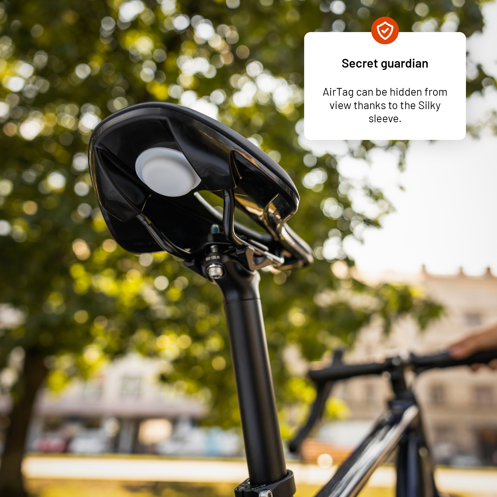 Ein Fahrradsattel mit weißer Scheibe, vermutlich ein AirTag, ist an einer schwarzen Sattelstütze befestigt. Der Hintergrund sind verschwommene grüne Bäume.