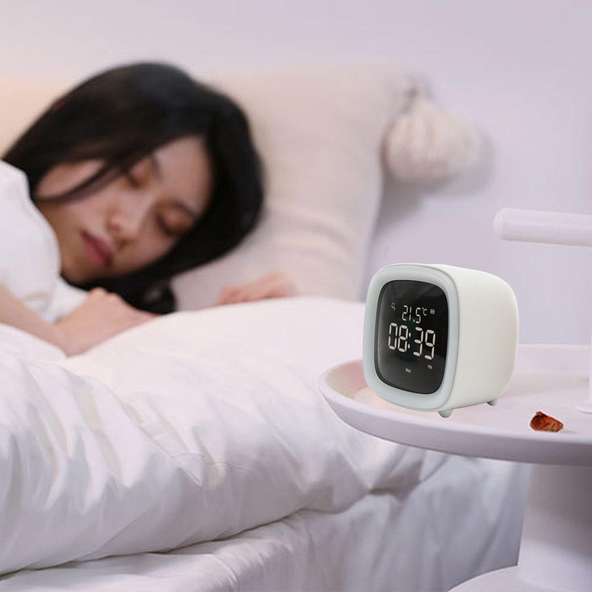 Eine Frau schläft im Bett. Eine weiße Uhr zeigt die Zeit an. Die Uhr steht auf einem kleinen Tisch neben dem Bett.