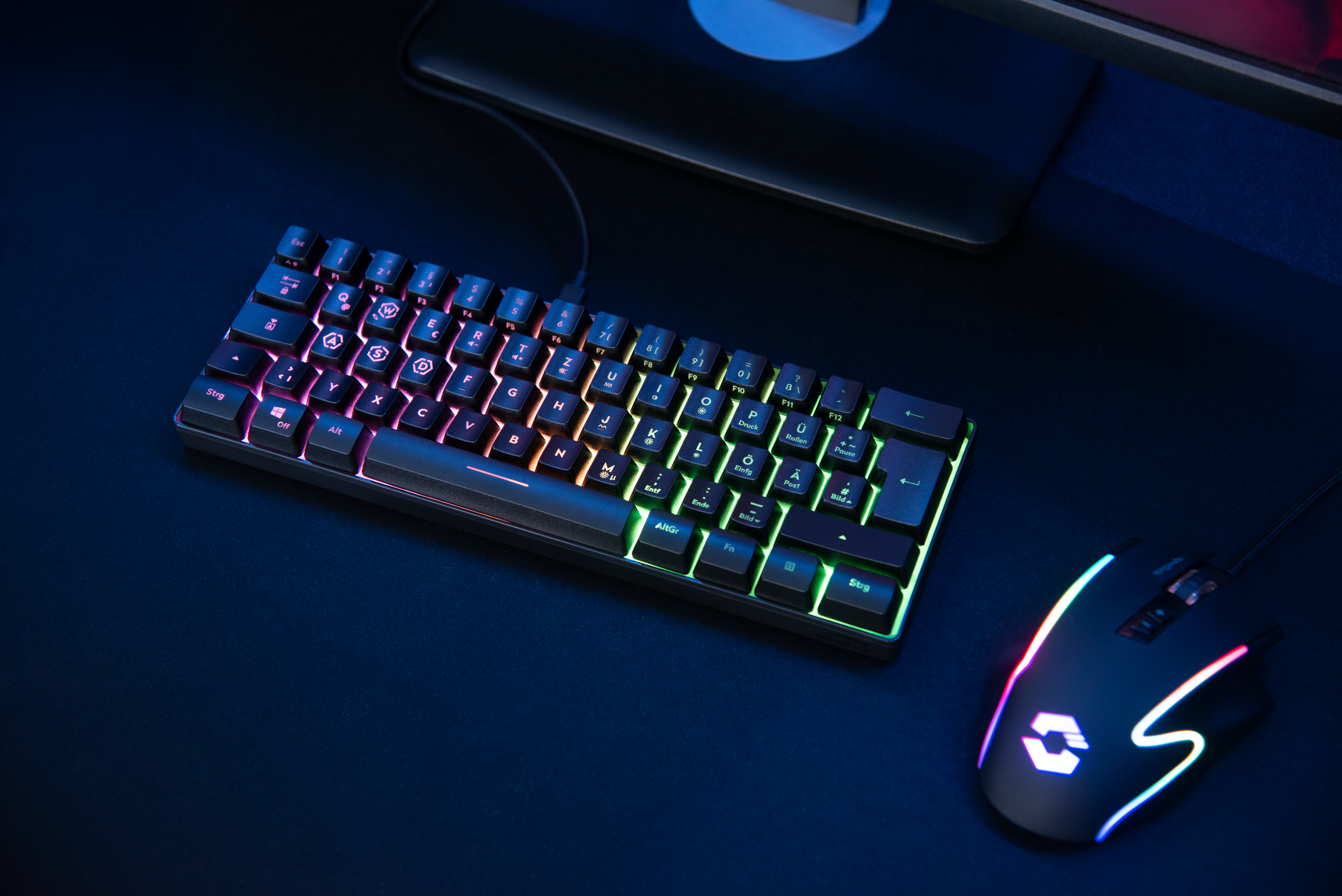 Gaming-Tastatur und -Maus mit bunten Lichtern auf dunkler Oberfläche, Monitor im Hintergrund.