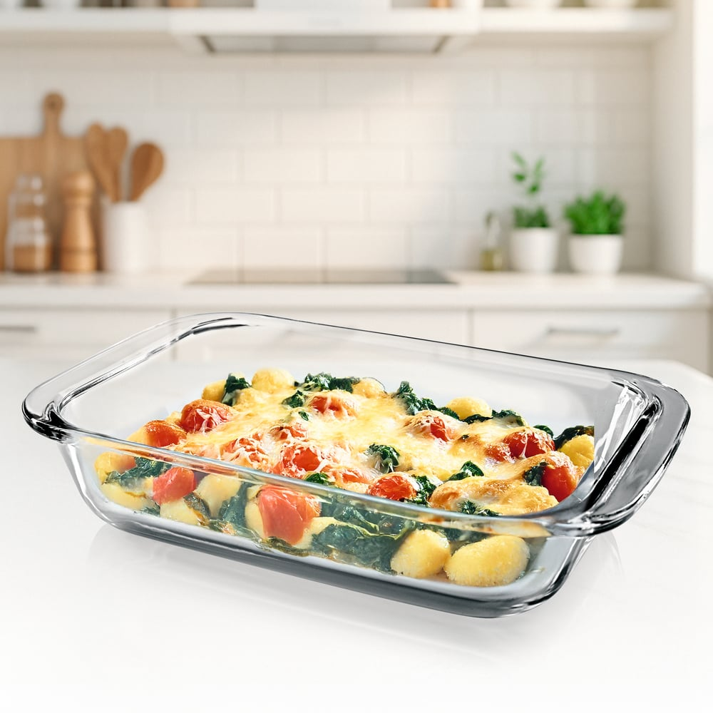 Eine Glasschale enthält gebackenes Essen: Tomaten, Spinat und Käse. Die Schale steht auf einer weißen Oberfläche. Küchenhintergrund.