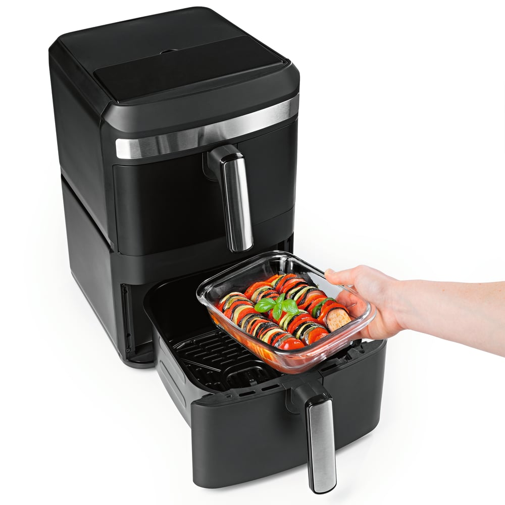 Ein schwarzer Airfryer mit einem Glasbehälter mit gekochtem Gemüse, das hineingelegt wird. Eine Hand hält die Schale mit Tomaten und Zucchini.