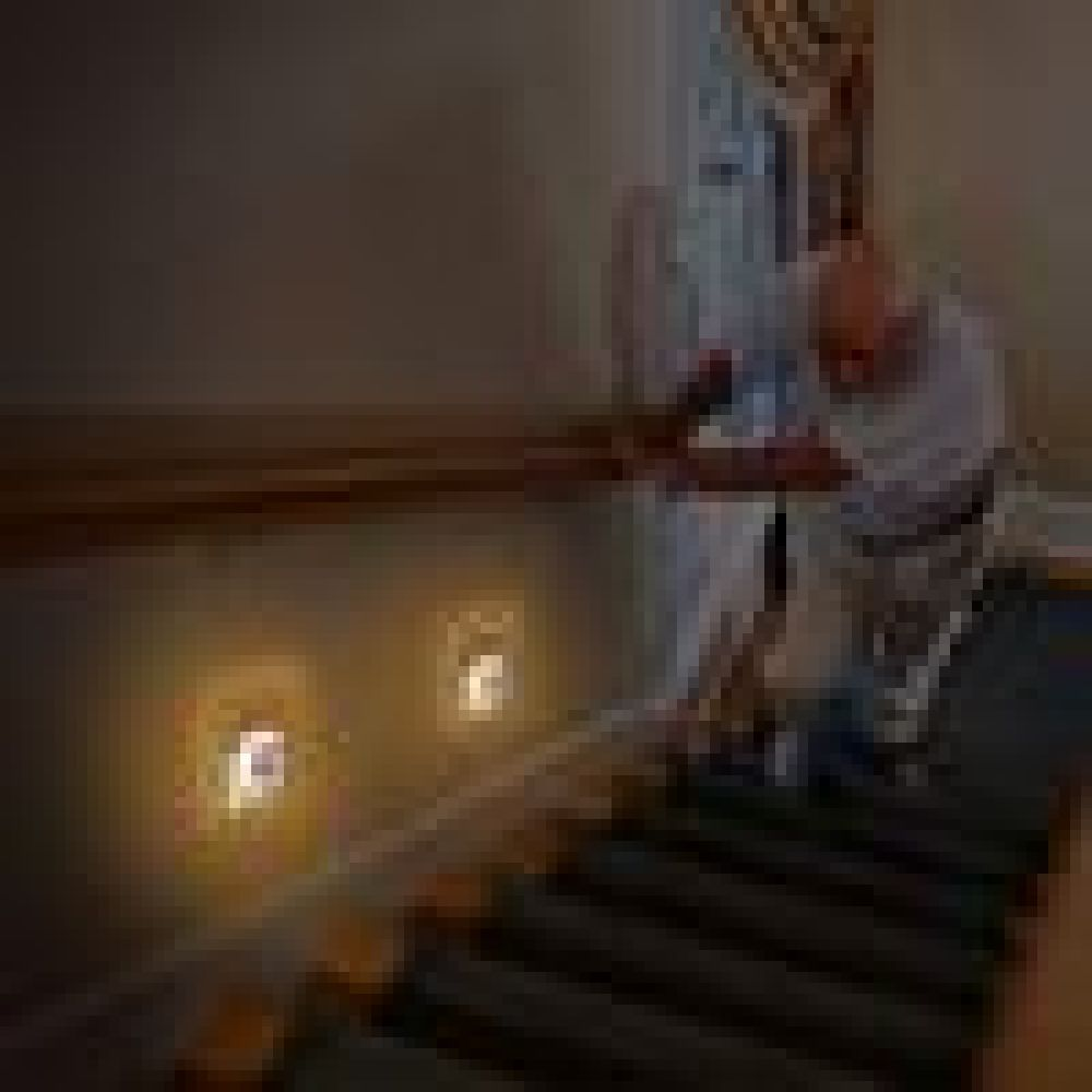Eine Person steht an einem Geländer und blickt auf eine Treppe. Zwei Lichter sind an der Wand zu sehen. Die Szene ist schwach beleuchtet.