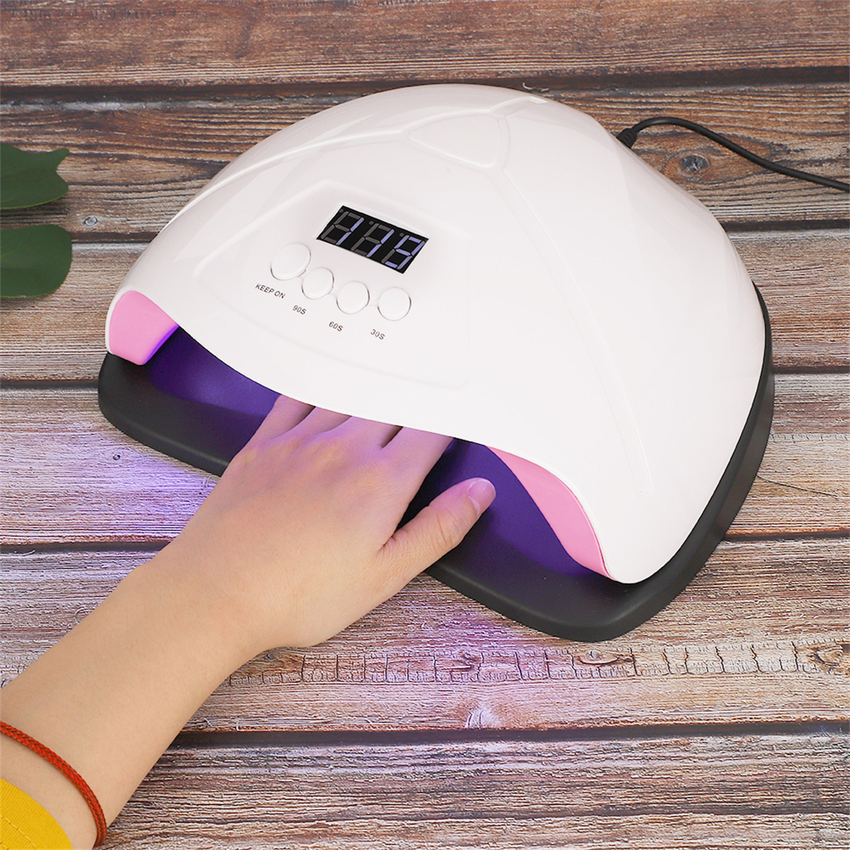 Eine Hand liegt in einer Nagelhärtungslampe. Die Lampe ist weiß und rosa. Digitalanzeige zeigt Zahlen. Holzoberfläche im Hintergrund.