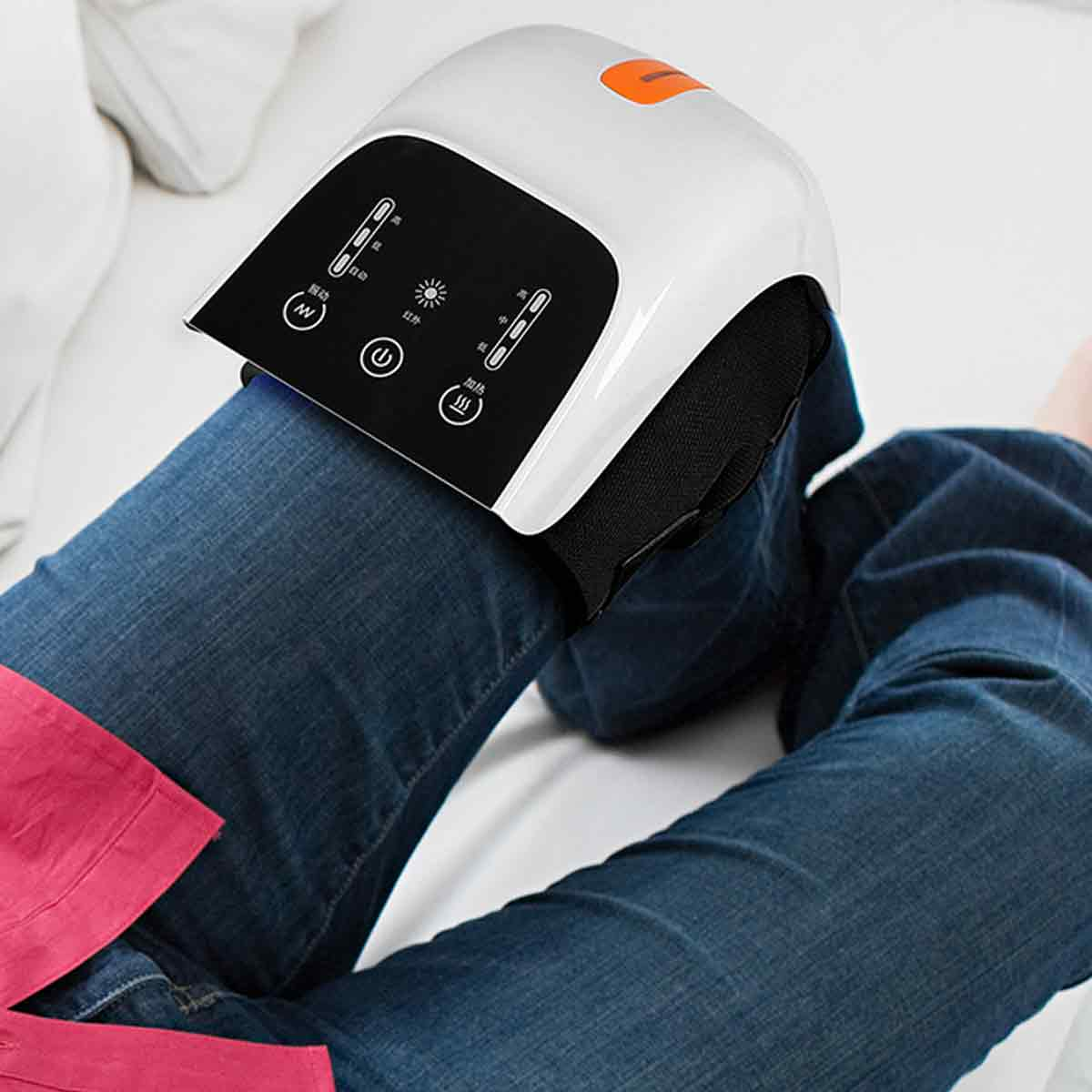 Eine Person mit Jeans hat ein Beinmassagegerät auf dem Knie. Das Massagegerät ist weiß und schwarz mit orangefarbenen Akzenten.
