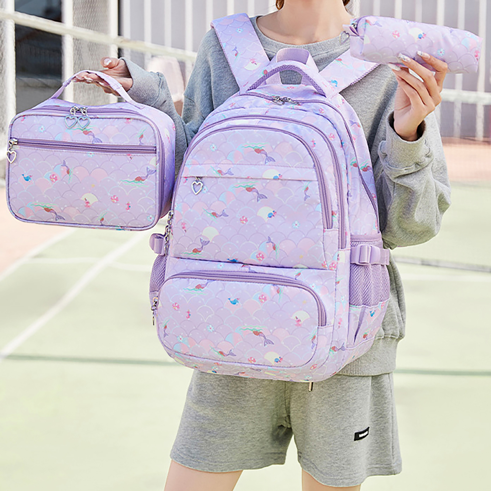 Eine Person hält einen passenden lila Rucksack, eine Lunchtasche und ein Federmäppchen. Das Set hat ein Meerjungfrauenmuster.