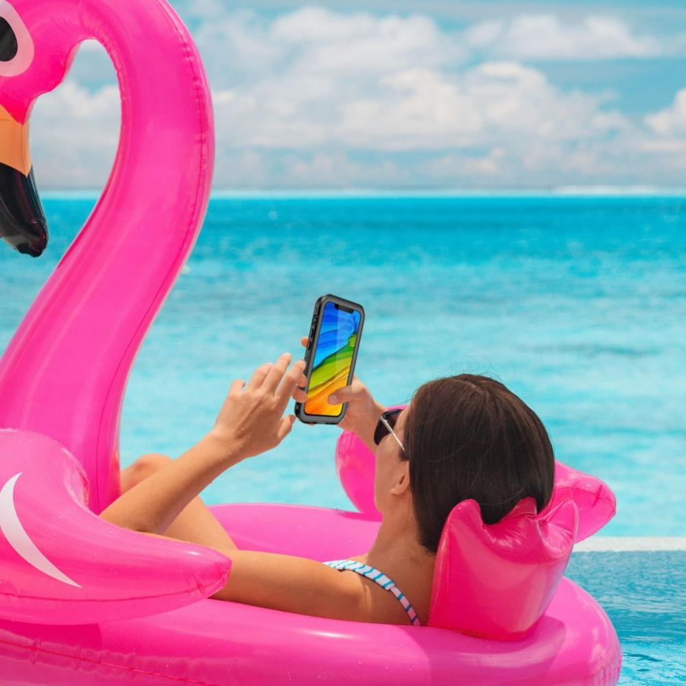 Eine Frau in einem Flamingo-Schwimmring schaut auf ihr Handy. Das Telefon zeigt ein farbenfrohes Bild, und sie ist in einem Pool mit dem Meer im Hintergrund.