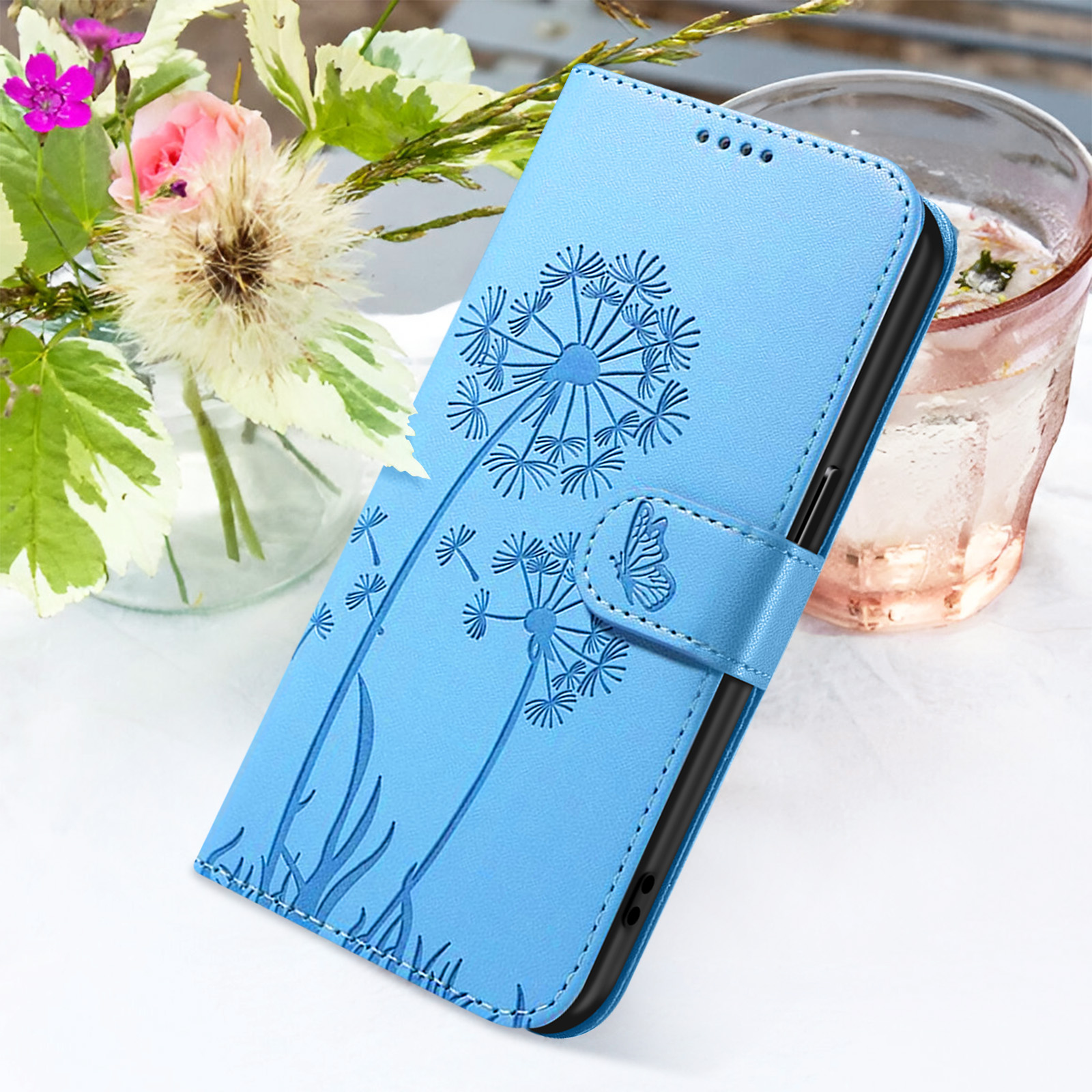 Una custodia blu per telefono con design a dente di leone è su una superficie bianca vicino a fiori e un bicchiere d'acqua.