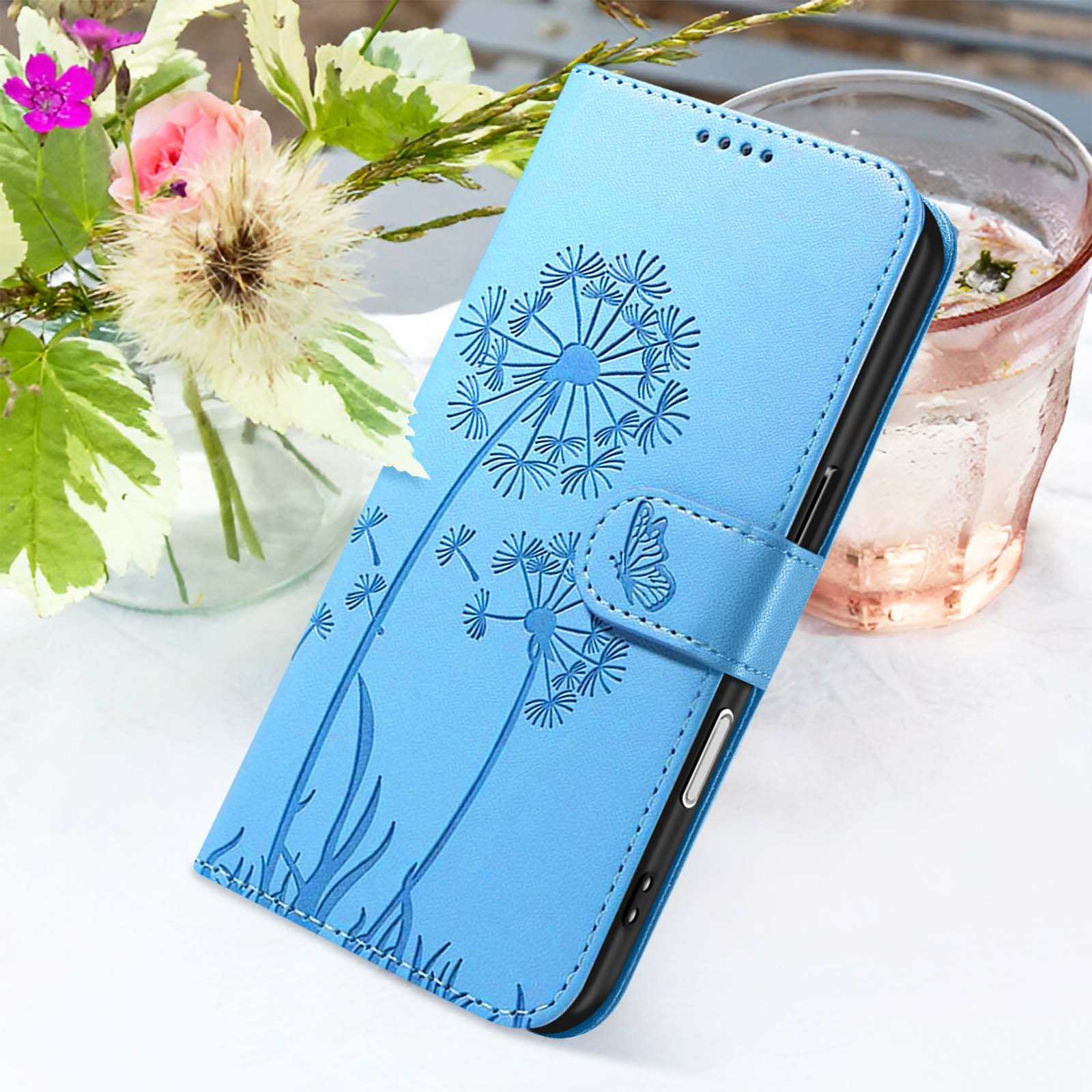 Una custodia blu per telefono con design a dente di leone è su una superficie bianca con fiori e un bicchiere d'acqua sullo sfondo.