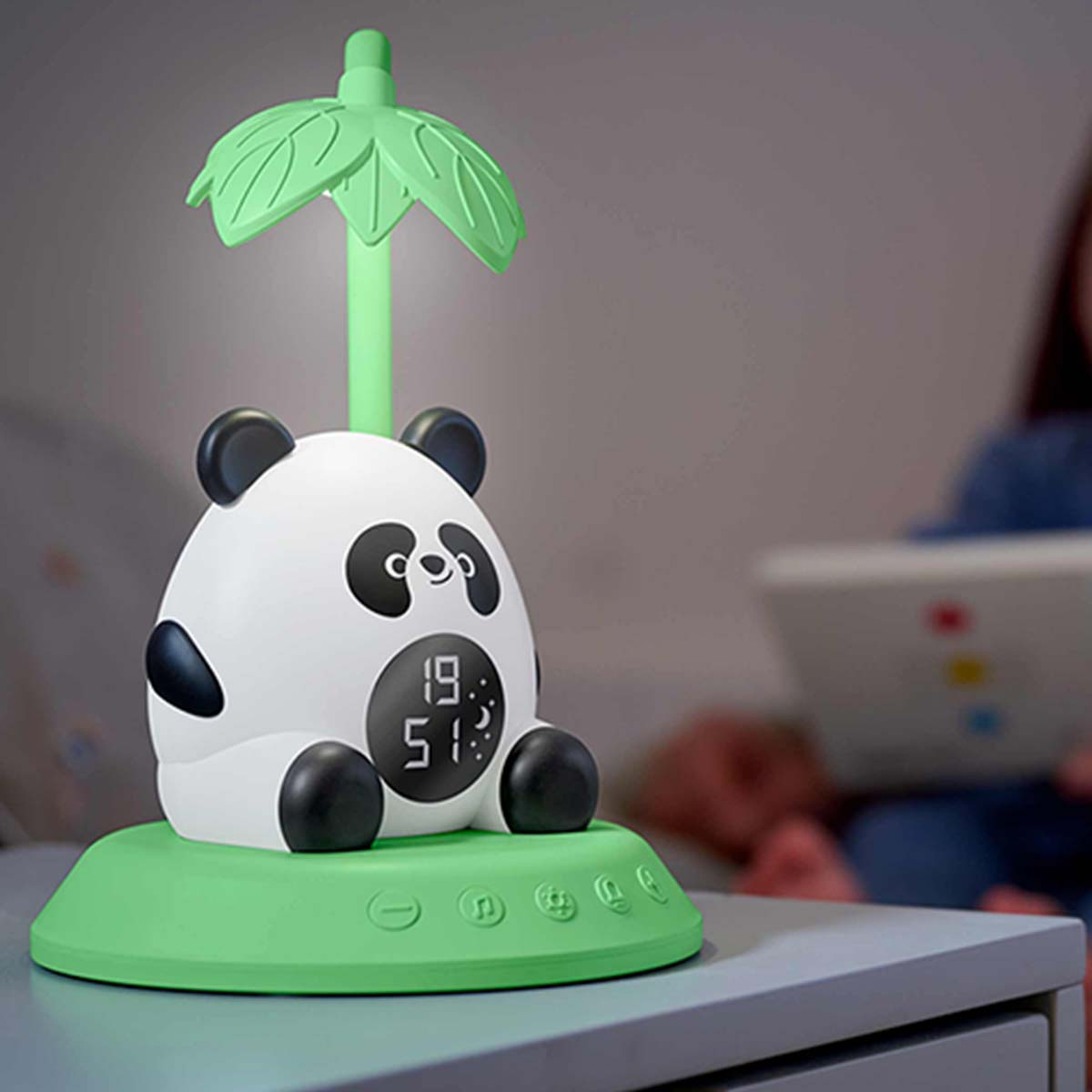 Una luce notturna a forma di panda con base verde e luce a forma di foglia. Una persona legge sullo sfondo.