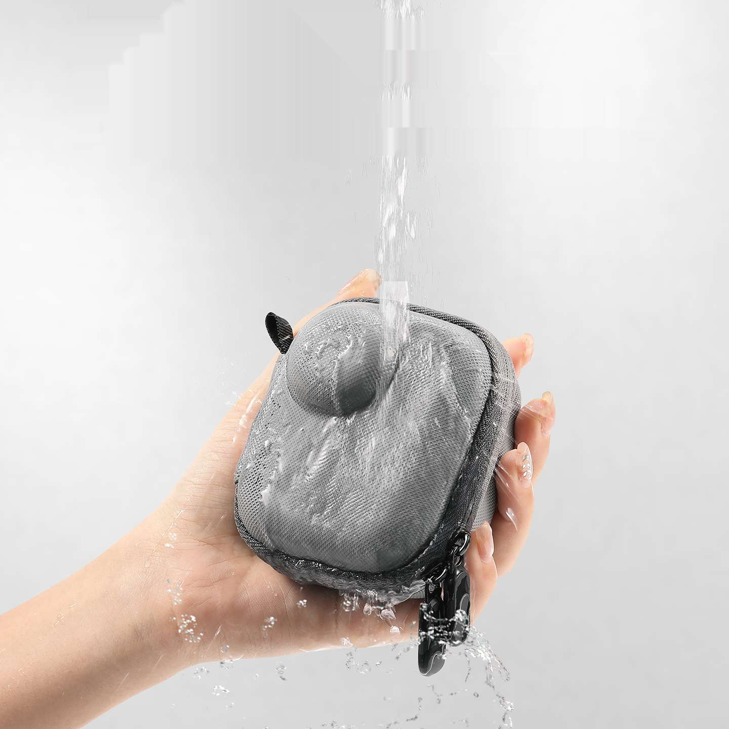 Eine Hand hält einen grauen, quadratischen Beutel. Wasser strömt auf den Beutel. Der Hintergrund ist weiß. Schwarzer Reißverschluss und kleine Schlaufe sind sichtbar.