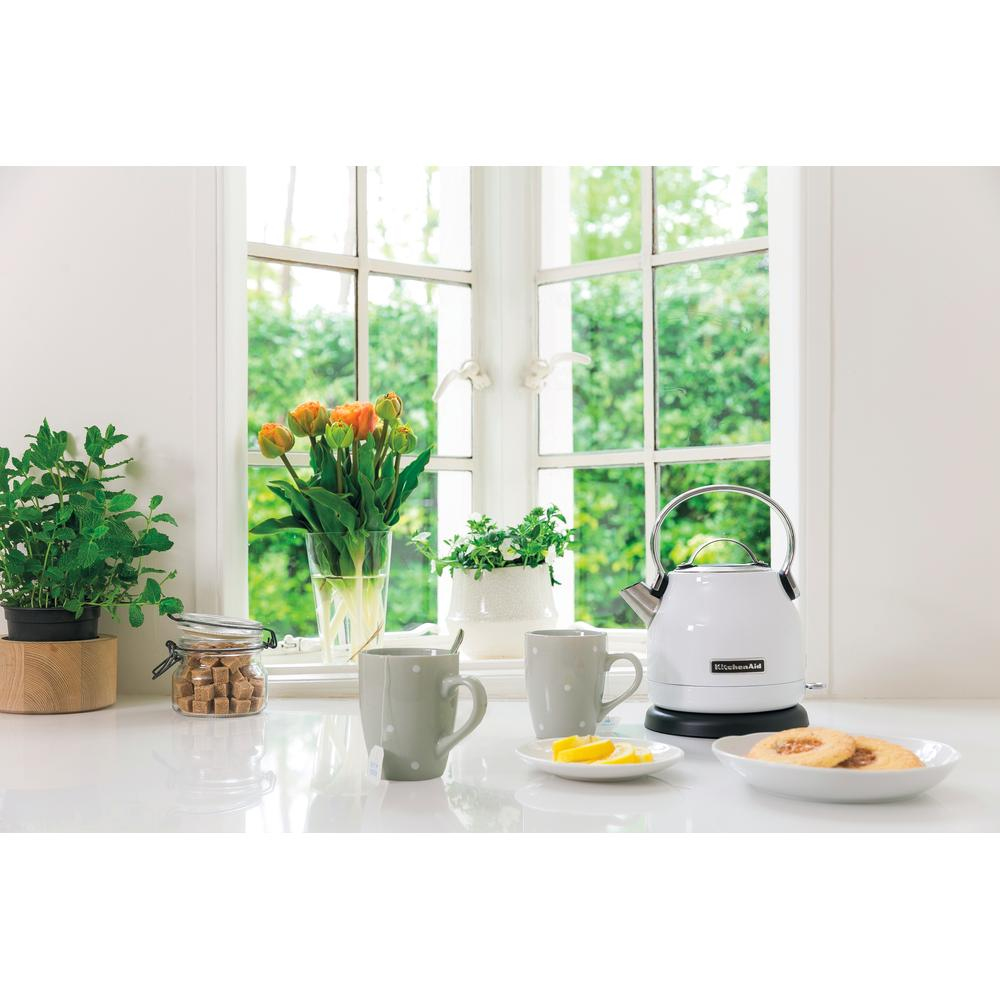 A kitchen scene with a white kettle, mugs, flowers, lemon slices, and cookies on a white countertop by a window.