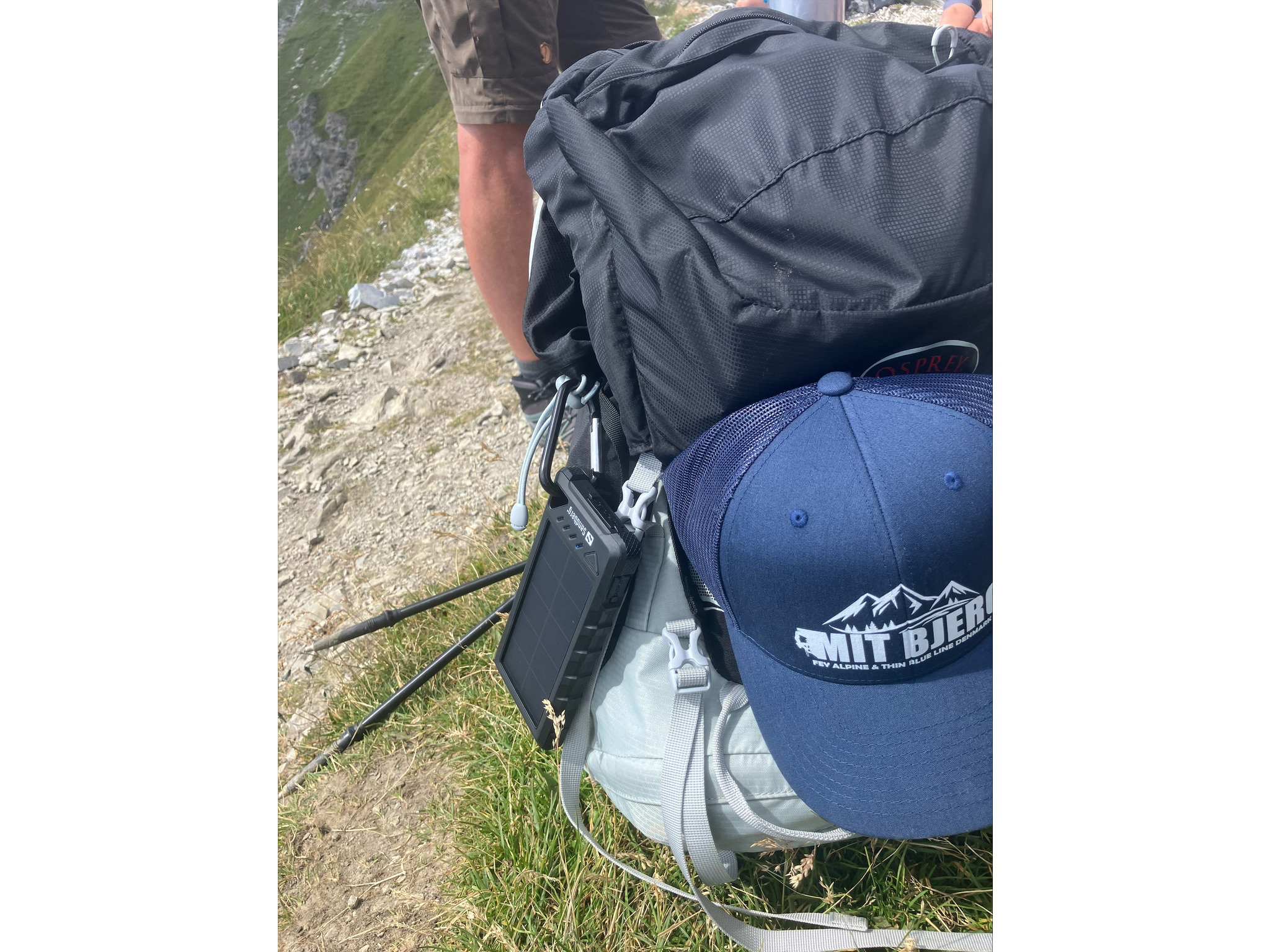 Ein Rucksack mit blauer Mütze und Solar Ladegerät. Wanderstöcke sind sichtbar. Der Hintergrund zeigt einen Bergpfad.