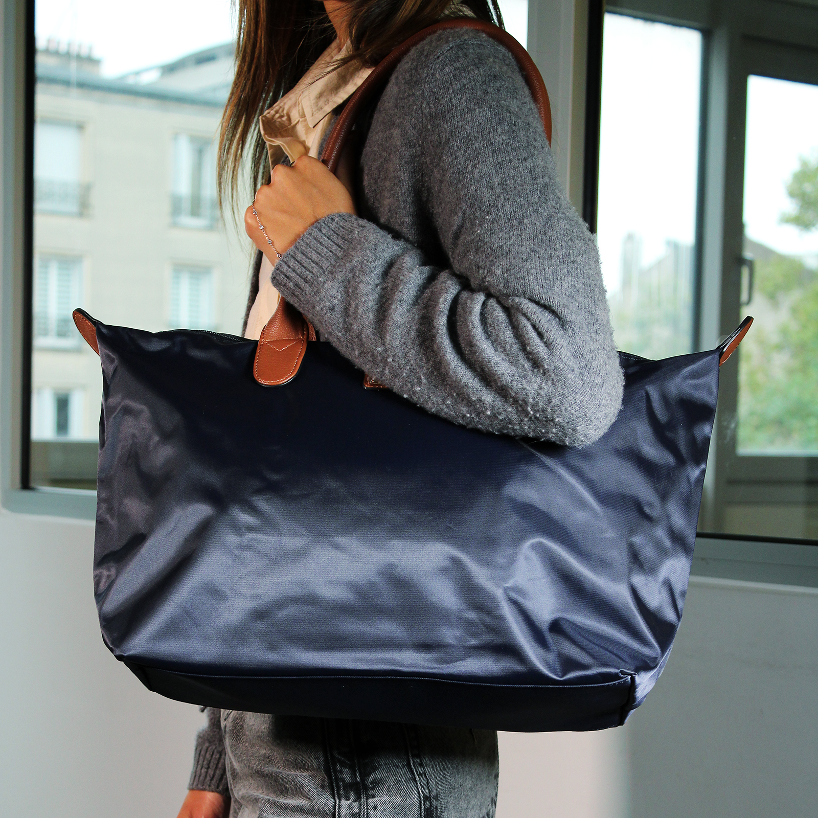 Una mujer sostiene un bolso tote azul marino con asas marrones. Lleva un suéter gris y está cerca de una ventana.