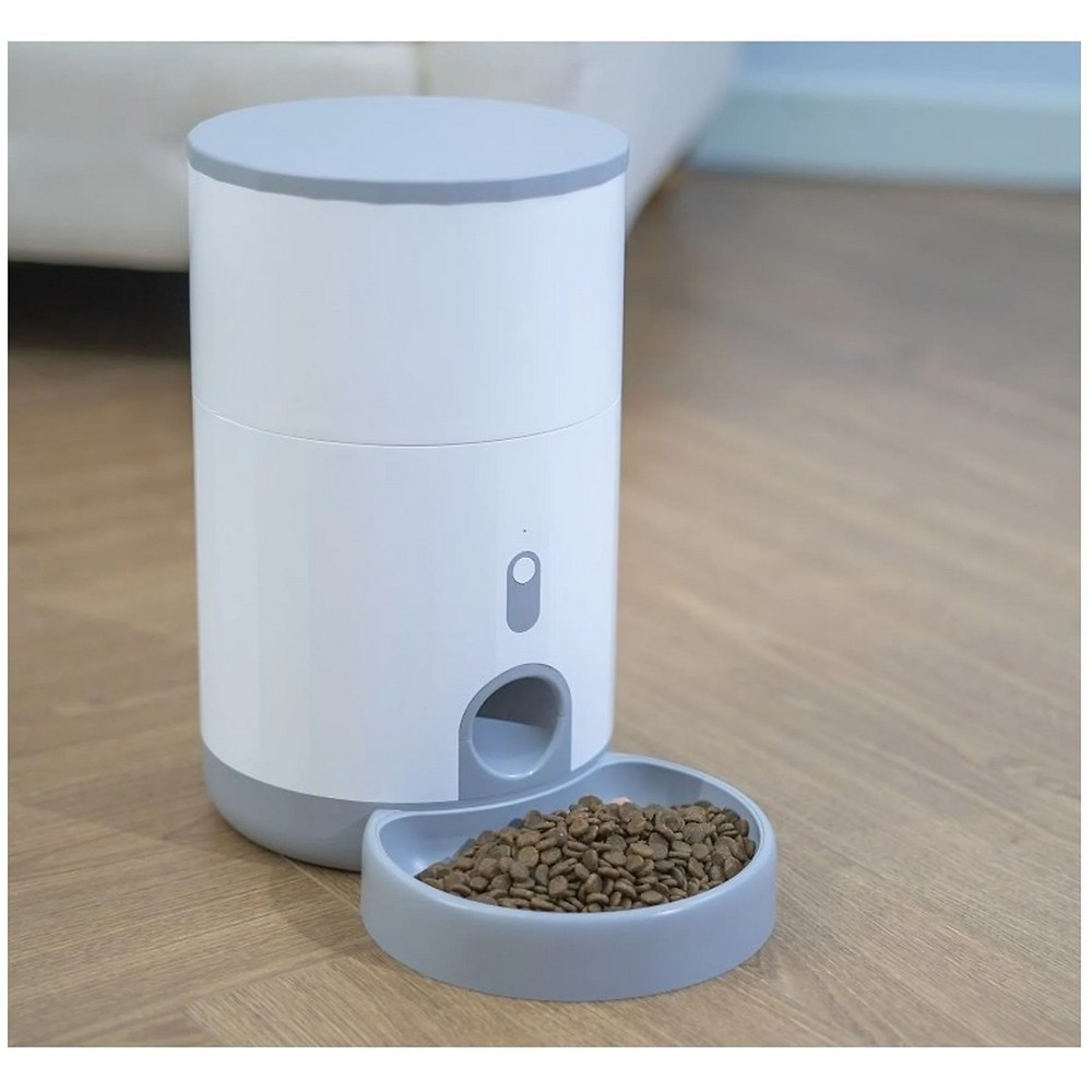 Biało-szary automatyczny podajnik karmy ze jedzeniem w misce. Stoi na drewnianej podłodze, z rozmytym tłem.