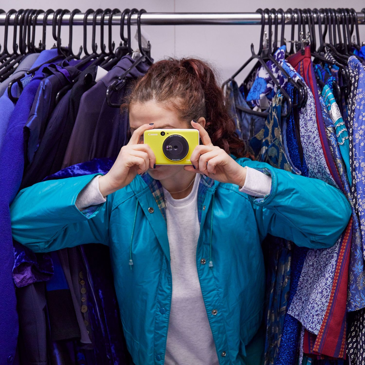 Una persona pelirroja sostiene una cámara amarilla, tomando una foto de ropa en un perchero. Lleva una chaqueta azul.