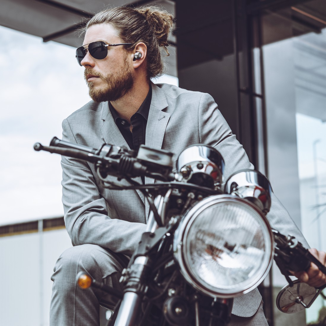 Mężczyzna w garniturze i okularach przeciwsłonecznych siedzi na motocyklu. Ma słuchawki. Motocykl jest czarny i srebrny.
