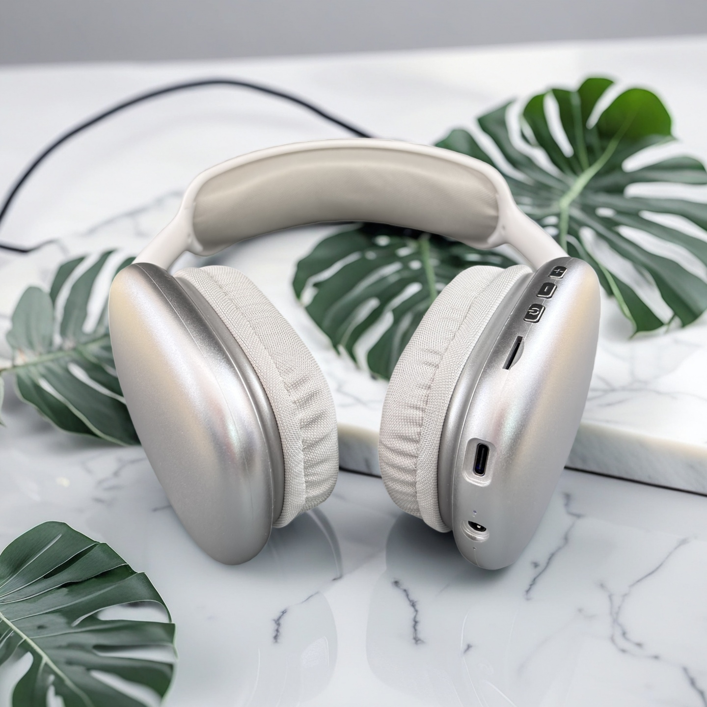 Auriculares plateados sobre mármol blanco, rodeados de hojas verdes. Los auriculares tienen orejeras gris claro y diadema blanca.