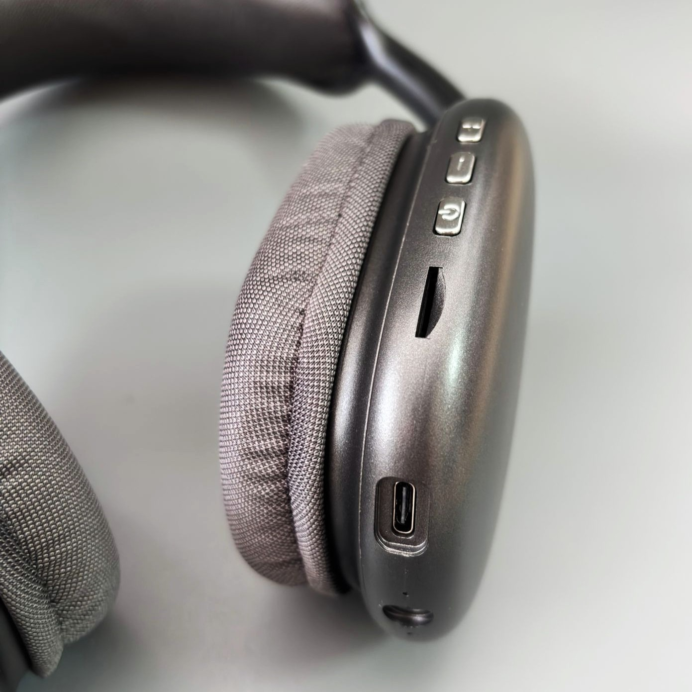 Primer plano de auriculares grises. El auricular derecho muestra botones, un puerto de carga y una ranura para tarjeta de memoria. El fondo es gris claro.