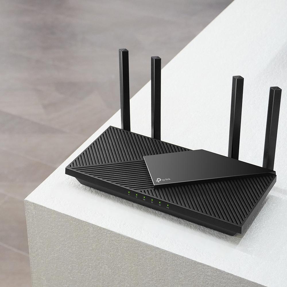 Czarny router TP-Link z czterema antenami stoi na białej powierzchni. Router ma płaską, kątową konstrukcję. Widoczne są zielone kontrolki.