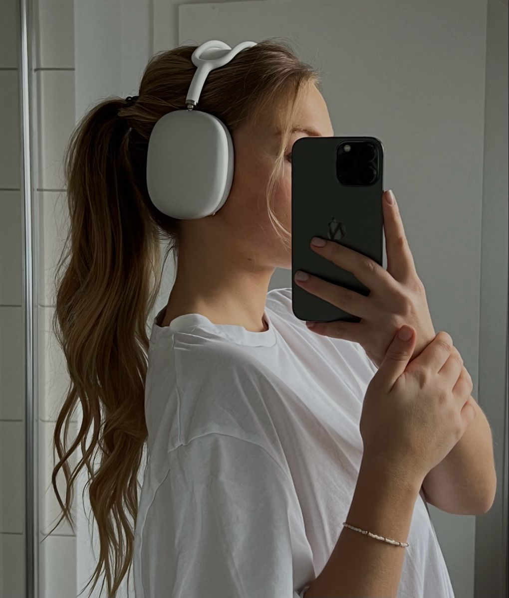 Mujer con camisa blanca y auriculares tomándose una selfie en el espejo con un teléfono. Pelo largo y rubio en cola de caballo.