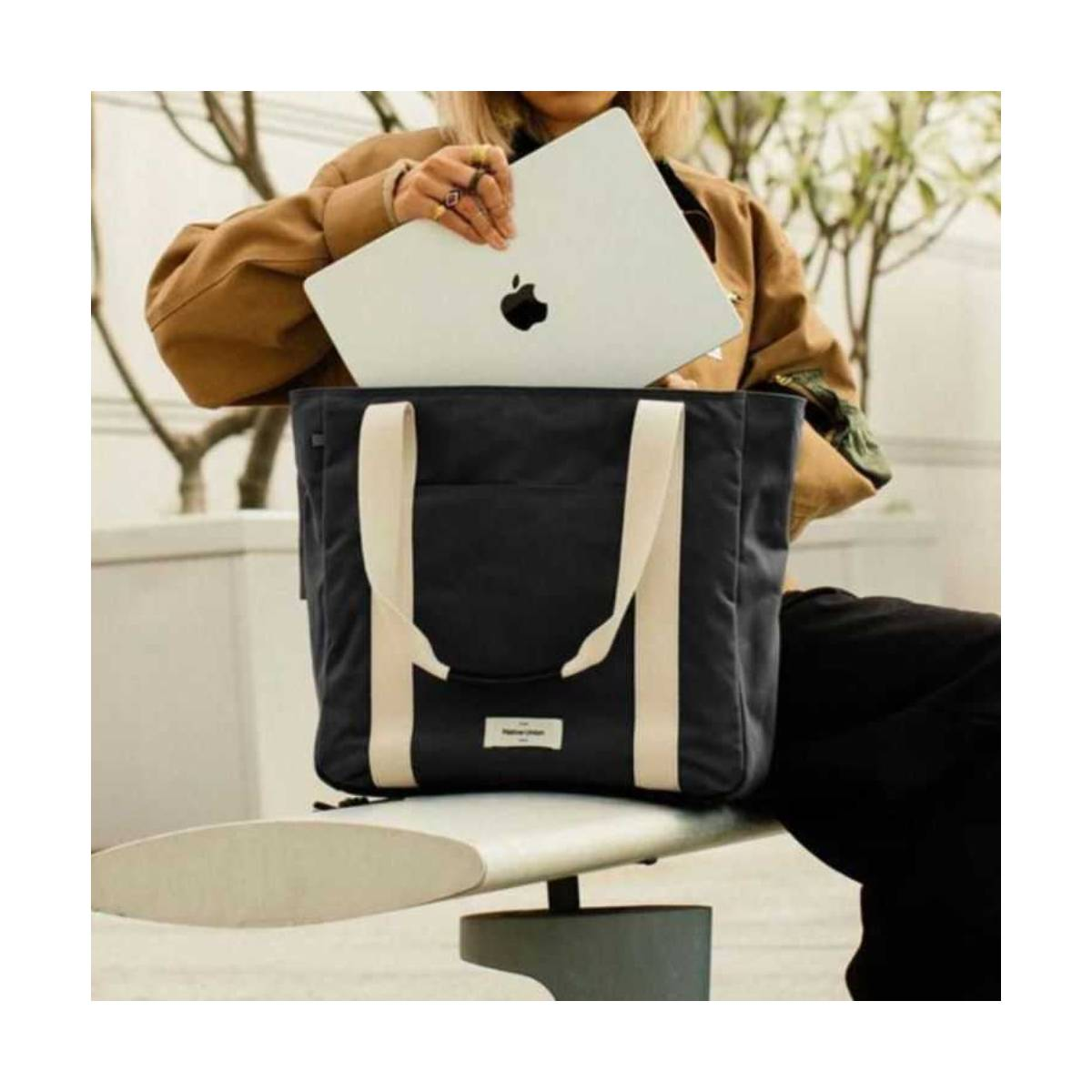 Una persona tiene un laptop argentato sopra una borsa tote nera con cinghie color crema, su una panca bianca. La persona indossa una giacca marrone.