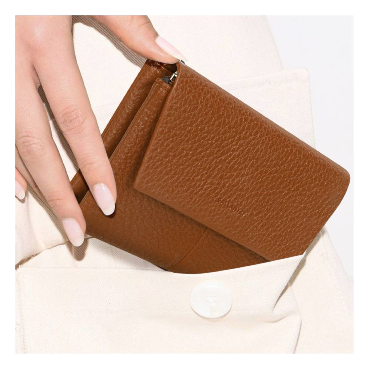 Una mano sostiene una cartera de cuero marrón. La cartera está parcialmente en un bolsillo de tela blanca. Las uñas están pintadas de blanco.