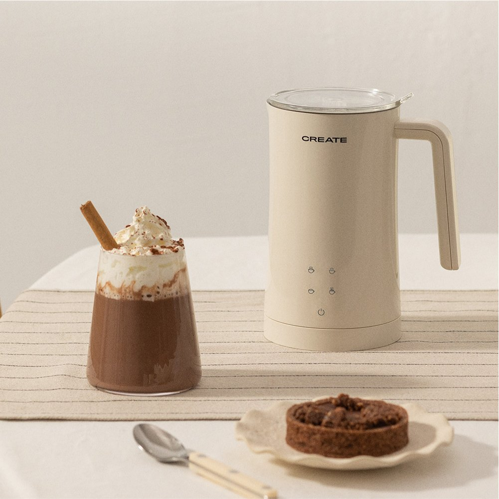 Una mesa con un vaso de bebida de chocolate, un espumador, una tarta y una cuchara. La bebida tiene crema batida y una rama de canela.