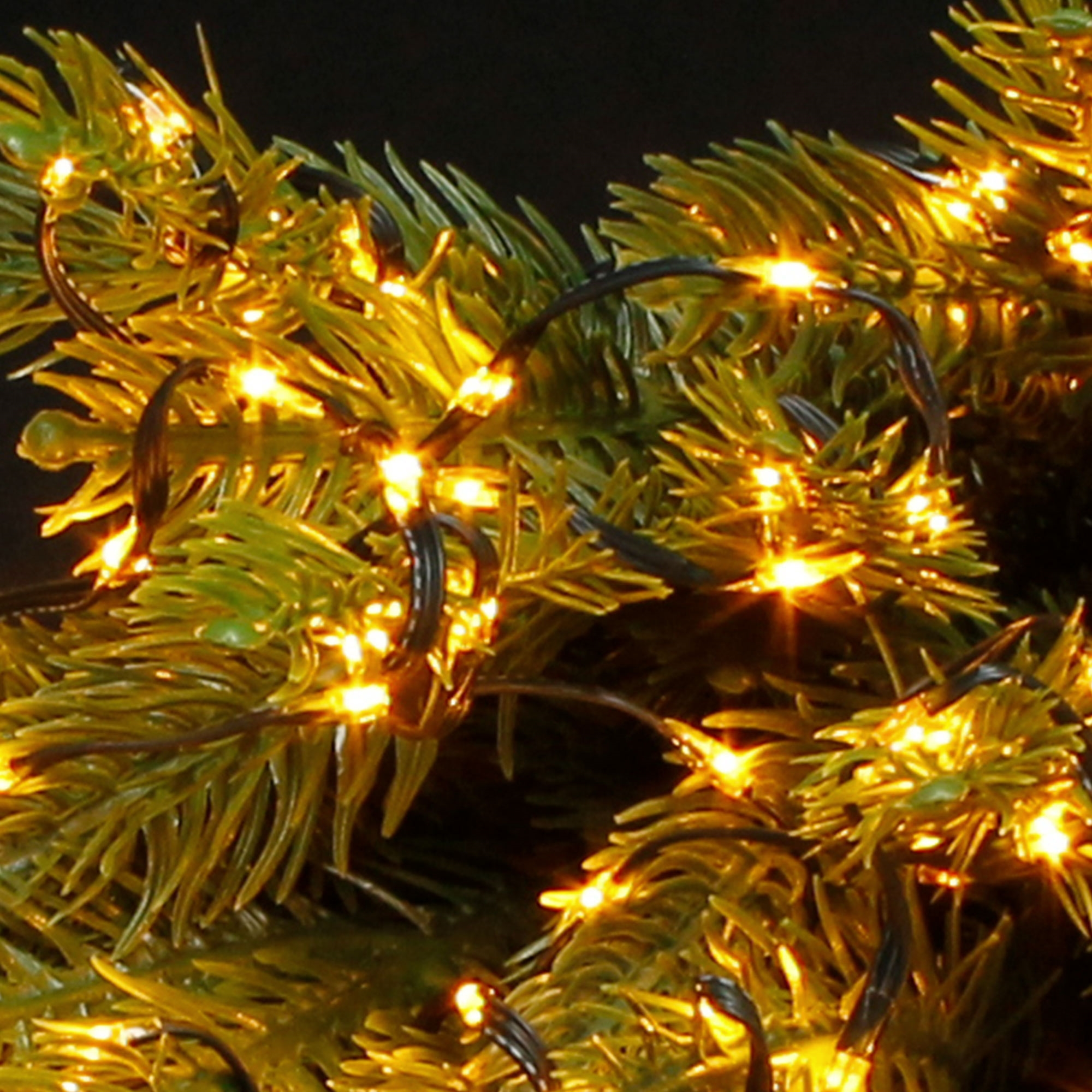 Nahaufnahme eines Weihnachtsbaums mit gelb leuchtenden Lichtern. Grüne Nadeln und schwarze Drähte sind sichtbar.