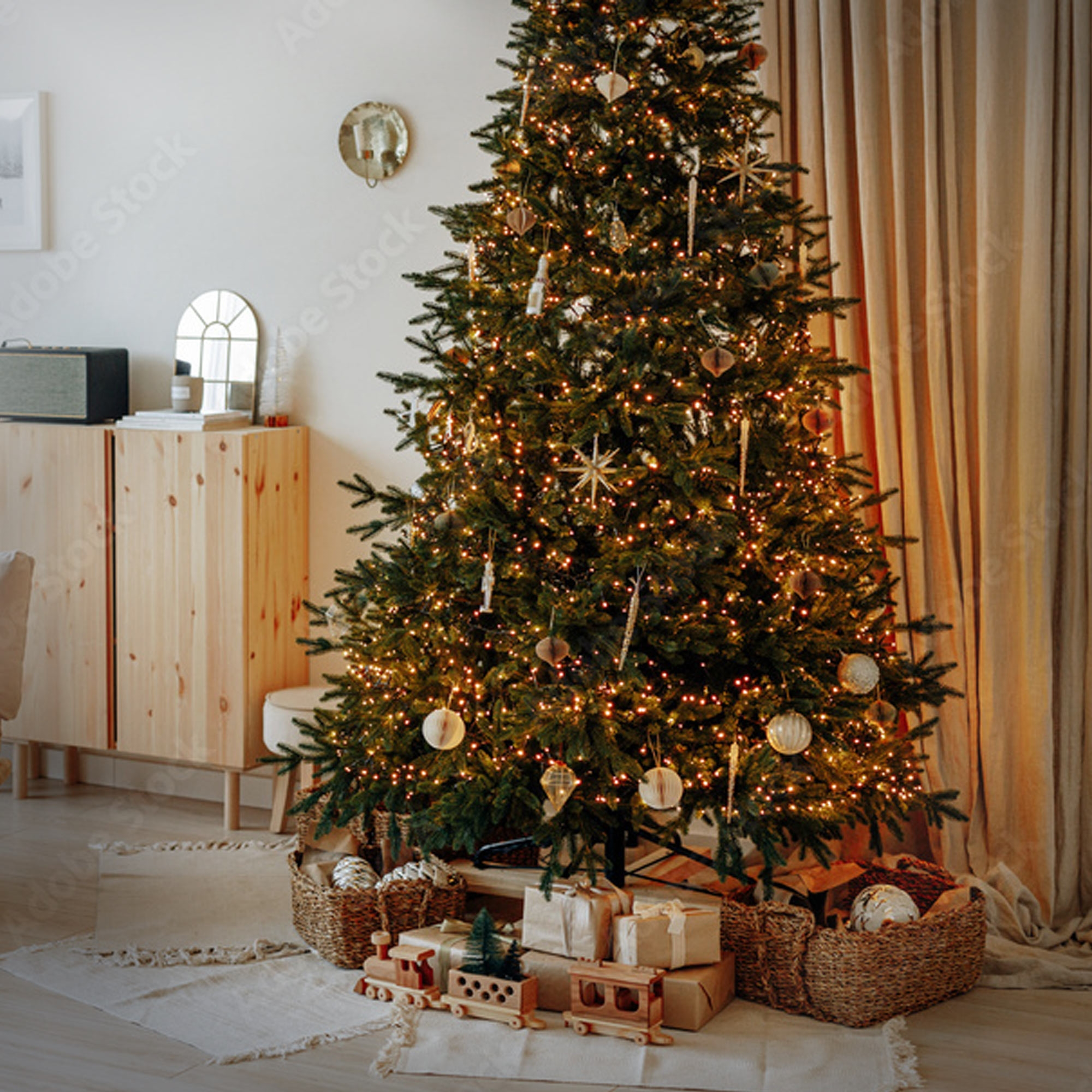Ein geschmückter Weihnachtsbaum mit Lichtern steht in einem Raum. Geschenke, Körbe und ein Holzzug befinden sich am Fuße des Baumes.