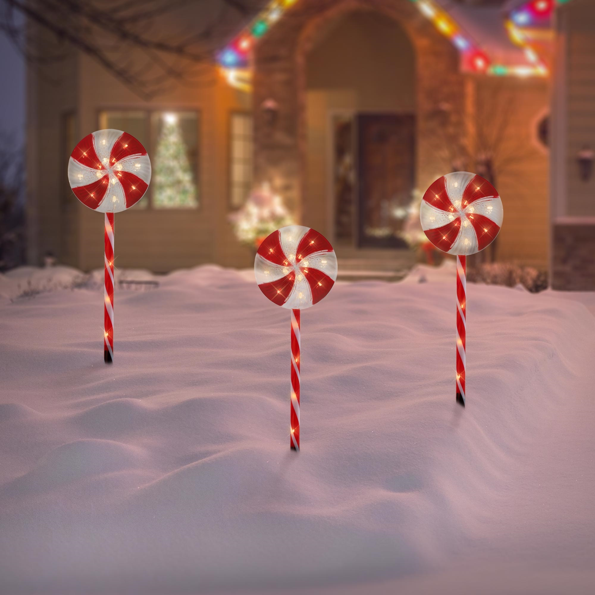Drei beleuchtete Zuckerstangen-Dekorationen im Schnee. Im Hintergrund ein Haus mit Weihnachtsbeleuchtung.