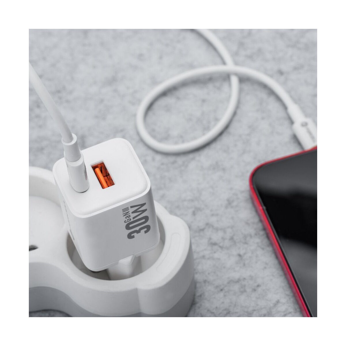 Un caricatore bianco da 30 W è collegato a una presa, collegato a uno smartphone rosso con un cavo bianco su una superficie grigia.